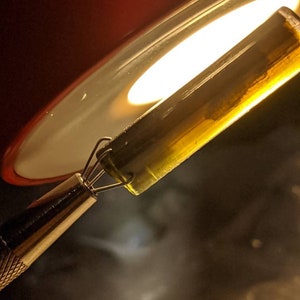 May include: A close-up of a clear glass tube filled with a yellow liquid. The tube is being held by a pair of tweezers and is being heated by a light source.