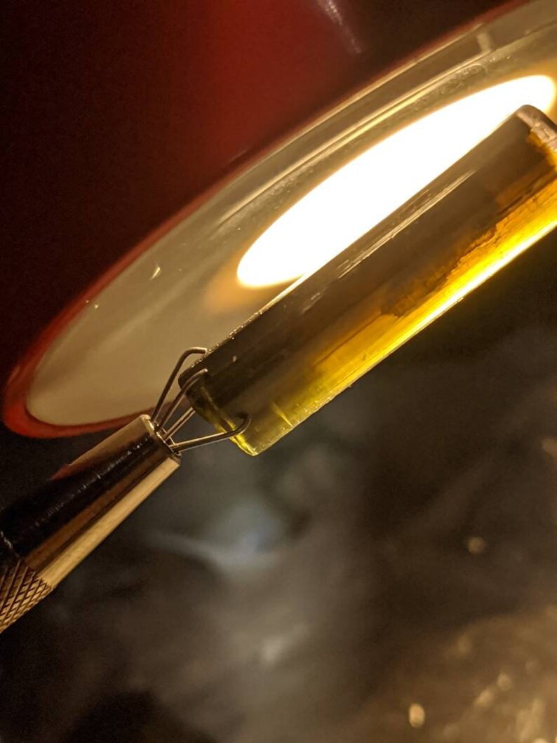 May include: A close-up of a clear glass tube filled with a yellow liquid. The tube is being held by a pair of tweezers and is being heated by a light source.