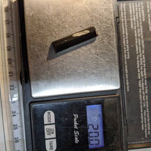 May include: A black digital pocket scale with a small black object on the weighing platform. The scale reads 2.00. The scale has buttons for mode, off, and tare.