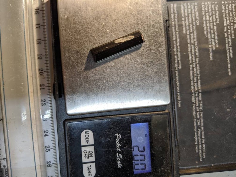 May include: A black digital pocket scale with a small black object on the weighing platform. The scale reads 2.00. The scale has buttons for mode, off, and tare.