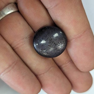 May include: A round, dark gray gemstone with a shimmery, iridescent finish. The gemstone is approximately 1 inch in diameter.