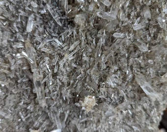 Puede incluir: Un cúmulo de cristales de cuarzo transparentes con una superficie rugosa y gris. Los cristales son largos y delgados, y están dispuestos en un patrón aleatorio. Los cristales son muy brillantes y reflejan la luz.