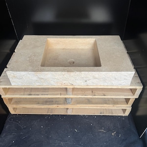 Puede incluir: Un lavabo rectangular de piedra de color claro con una cubeta cuadrada. El lavabo está colocado sobre un palet de madera. El fondo es negro.