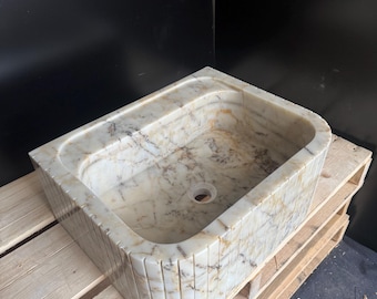 May include: A rectangular marble sink with a rounded corner. The sink is beige with brown veining and a drain hole. The exterior has vertical grooves. It sits on a wooden pallet against a black background.