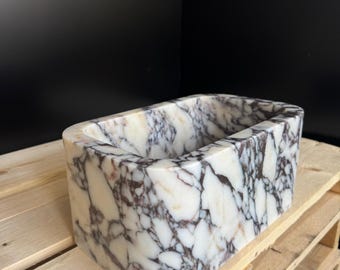 May include: A rectangular marble sink with rounded edges. The sink features a white base with intricate purple and gray veining. It is set on a wooden pallet against a black background. The sink's design is modern and elegant.