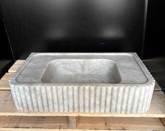 May include: A rectangular, white marble sink with a fluted exterior. The sink has a recessed basin and a flat, square rim. The marble has subtle gray veining, and the sink is displayed on a wooden pallet.