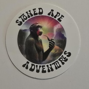 May include: Round sticker with the text "STONED APE ADVENTURES" in black, surrounding a graphic of a monkey smoking a pipe. The monkey is in front of a colorful, psychedelic background.