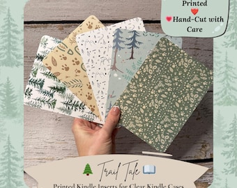 May include: A hand holds a fan of printed Kindle inserts with various nature-themed designs. The inserts feature patterns of trees, paw prints, and mountain scenes. Text on the image reads "Trail Tale" and "Printed Kindle Inserts for Clear Kindle Cases."