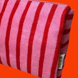 May include: A rectangular zippered pouch made of a soft, textured fabric featuring vertical stripes in alternating shades of bright pink and deep red. A small white fabric tag with gold-colored text is attached to the side seam of the pouch.