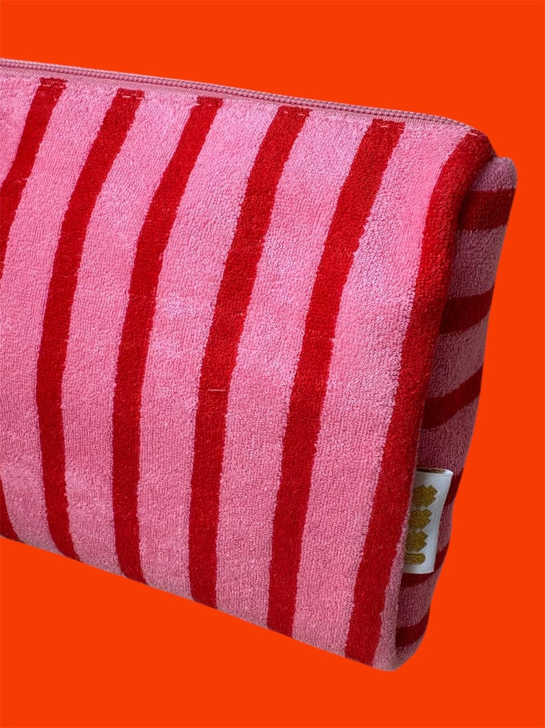 May include: A rectangular zippered pouch made of a soft, textured fabric featuring vertical stripes in alternating shades of bright pink and deep red. A small white fabric tag with gold-colored text is attached to the side seam of the pouch.