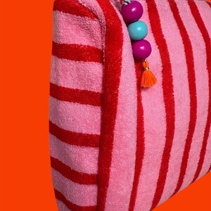 May include: A zippered pouch made of pink and red striped terry cloth fabric. The silver metal zipper pull is decorated with two purple wooden beads, one light blue wooden bead, and a small orange tassel hanging from the end of the zipper.