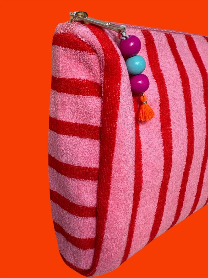 May include: A zippered pouch made of pink and red striped terry cloth fabric. The silver metal zipper pull is decorated with two purple wooden beads, one light blue wooden bead, and a small orange tassel hanging from the end of the zipper.