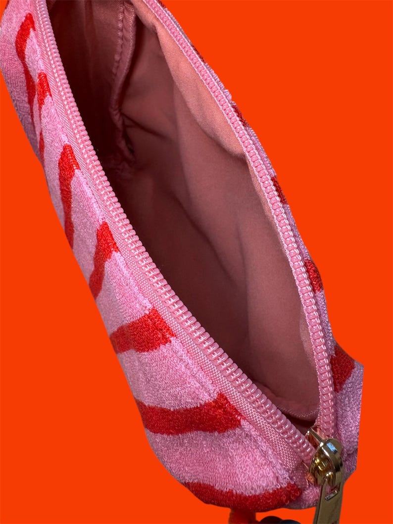 May include: A small zippered pouch made of a textured, terry cloth-like fabric featuring horizontal stripes in alternating shades of bright pink and vibrant red. The interior is lined with a solid light pink fabric and includes a pink zipper closure.