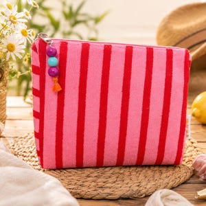 May include: A rectangular zippered pouch featuring vertical stripes in alternating shades of bright pink and red. The zipper pull is decorated with three round beads in purple and blue, finished with a small orange tassel hanging from the end.