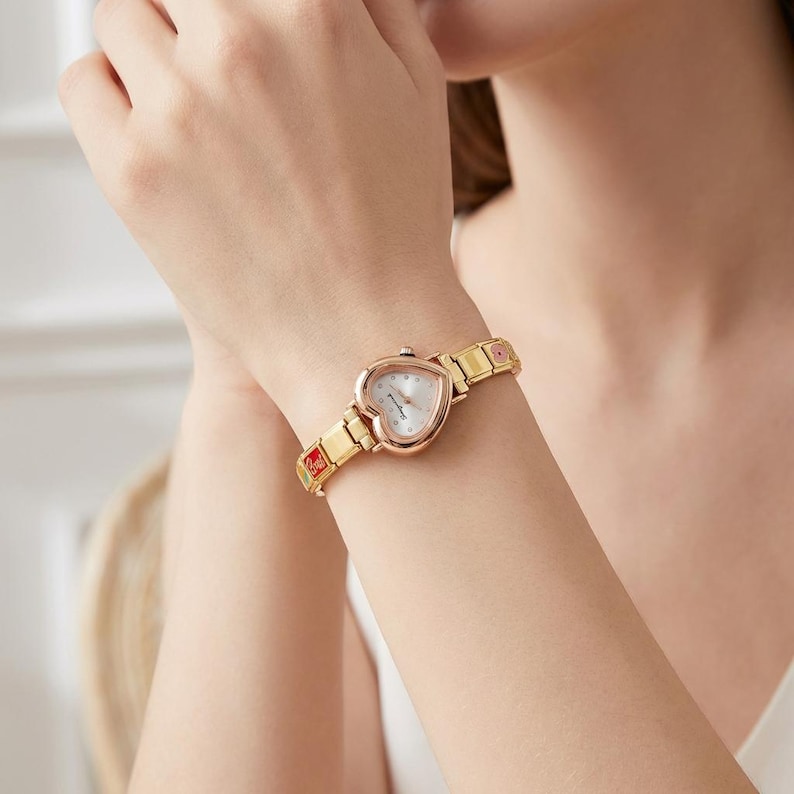 Puede incluir: Un reloj de pulsera dorado con una esfera en forma de coraz&oacute;n y una correa dorada. La esfera del reloj es blanca con agujas y marcadores dorados. La correa presenta eslabones decorativos con detalles rojos y rosas.