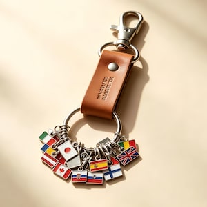 May include: A tan leather keychain featuring a silver metal lobster clasp and a large ring. Attached to the ring are multiple small, rectangular enamel charms, each displaying a different national flag, including Japan, Canada, Spain, and the UK.