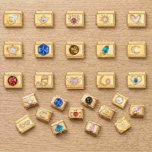 Può includere: Una collezione di braccialetti con ciondoli dorati con vari disegni, tra cui stelle, cuori, fiori e note musicali. Ogni ciondolo presenta una gemma o un elemento decorativo diverso, come una perla o un sole.