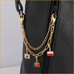 May include: A gold-toned metal chain accessory attached to a black textured bag. The accessory features two layered chains with lobster clasps and three rectangular enamel charms depicting the national flags of South Korea, Portugal, and Poland.