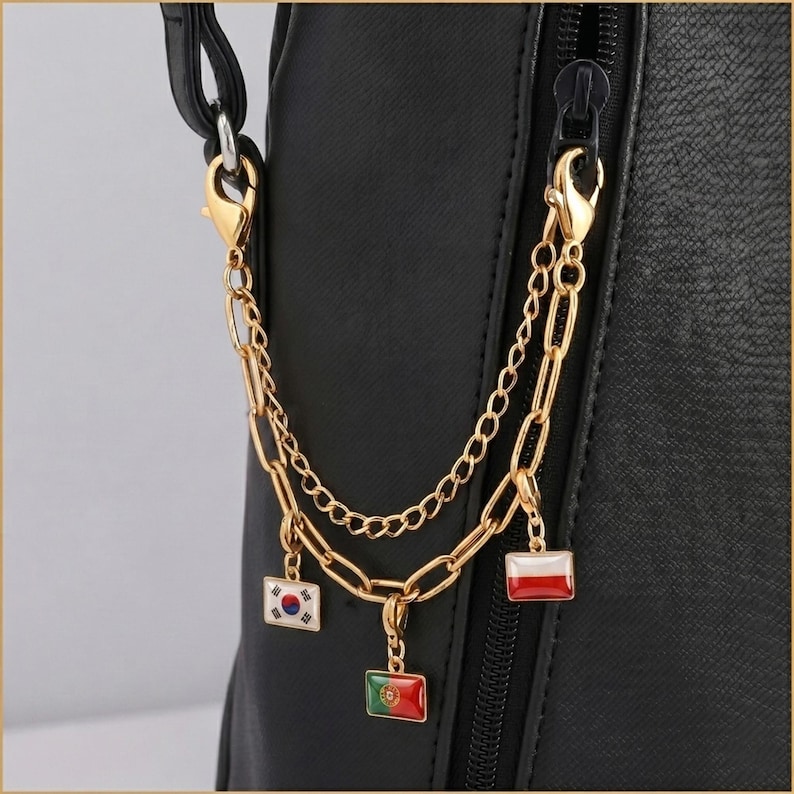 May include: A gold-toned metal chain accessory attached to a black textured bag. The accessory features two layered chains with lobster clasps and three rectangular enamel charms depicting the national flags of South Korea, Portugal, and Poland.