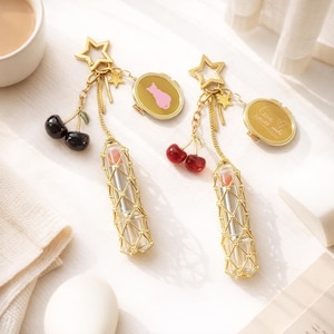 Könnte beinhalten: Zwei goldfarbene Lippenstifthalter mit Stern- und Kirschanhängern. Jeder Halter enthält einen Lippenstift und ein kleines goldenes Medaillon. Ein Medaillon zeigt eine rosa Katze, das andere lautet "Love never ends."