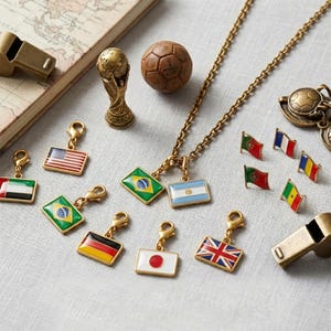 May include: A collection of gold-toned metal charms featuring various national flags, a miniature World Cup trophy figurine, a small brown soccer ball, and two whistles, arranged alongside a vintage-style map on a light-colored fabric surface.