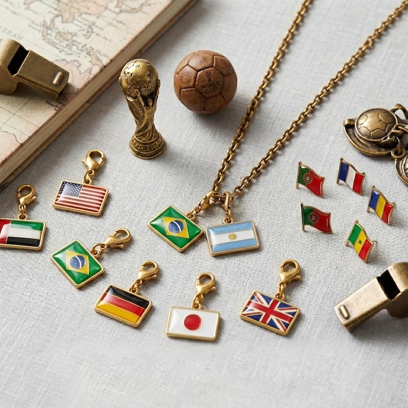 May include: A collection of gold-toned metal charms featuring various national flags, a miniature World Cup trophy figurine, a small brown soccer ball, and two whistles, arranged alongside a vintage-style map on a light-colored fabric surface.
