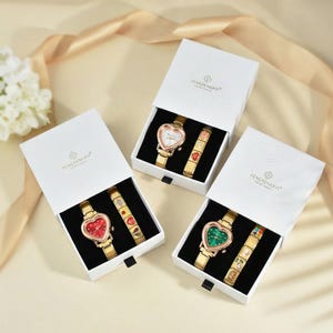 Puede incluir: Tres cajas blancas, cada una con un reloj dorado con esfera en forma de coraz&oacute;n y una pulsera a juego. Las esferas de los relojes son blancas, rojas y verdes. Las pulseras tienen elementos decorativos coloridos.