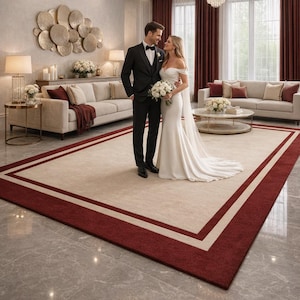 May include: A newly married couple in a luxurious room. The bride wears a white off-the-shoulder gown, and the groom is in a black tuxedo. They stand on a large beige rug with a burgundy border. The room features chandeliers and sofas.