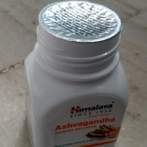 May include: A white plastic bottle of Himalaya Ashwagandha tablets. The bottle has a silver foil seal and a label with text including "Ashwagandha", "Pure Herbs", and "60 Tablets". The product is for general wellness.