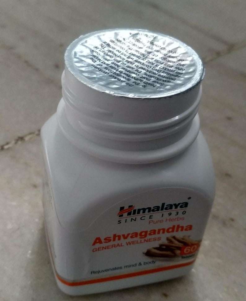 May include: A white plastic bottle of Himalaya Ashwagandha tablets. The bottle has a silver foil seal and a label with text including "Ashwagandha", "Pure Herbs", and "60 Tablets". The product is for general wellness.