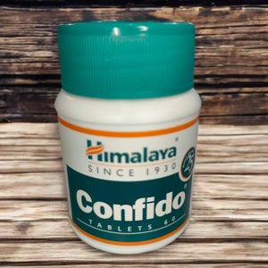 May include: A white and teal bottle of Himalaya Confido tablets. The bottle has a teal cap and a label with the Himalaya logo and the text "Confido Tablets 60". The background is a wooden surface.
