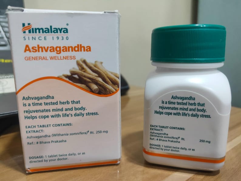 May include: A white box and bottle of Himalaya Ashvagandha supplements. The box features the brand logo and text, along with an illustration of the herb. The bottle has a green cap and white label with product information.