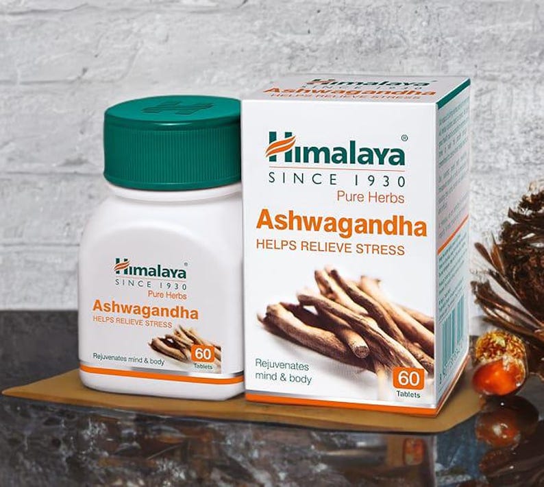 May include: A white bottle and box of Himalaya Ashwagandha tablets. The bottle has a green cap and the box displays the text "Helps Relieve Stress". The image shows the product on a wooden surface.