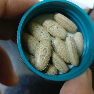 May include: A close-up shot of a teal-colored bottle filled with oval-shaped, light brown pills. The pills have a textured surface and are marked with the letters "AS". A white and orange box is partially visible in the background.