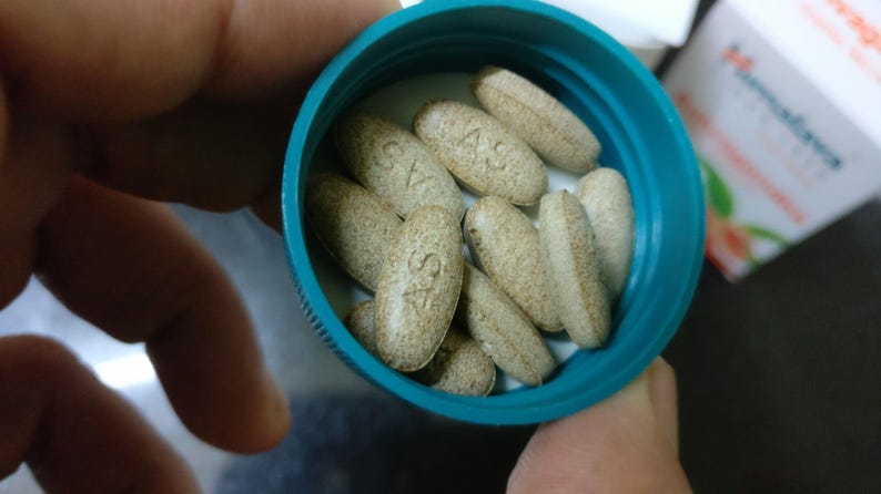 May include: A close-up shot of a teal-colored bottle filled with oval-shaped, light brown pills. The pills have a textured surface and are marked with the letters "AS". A white and orange box is partially visible in the background.
