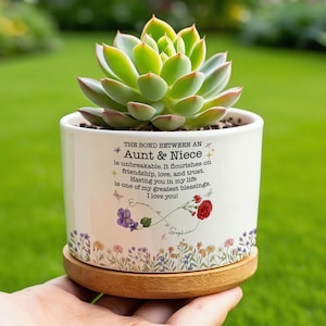 May include: A white ceramic planter with a succulent plant, featuring the text "THE BOND BETWEEN AN Aunt & Niece". The planter has a wooden base and is decorated with floral and butterfly designs. The succulent has green and red leaves.