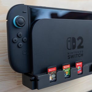 May include: Black Nintendo Switch console with attached controllers and game cartridges. The console is mounted on a black stand with the text "Nintendo Switch" and the number "2". The controllers have blue and orange accents.