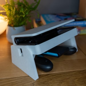 Peut inclure: Support blanc pour console de jeu avec une console noire et des manettes. Le support est posé sur une surface en bois, avec une petite plante en pot et une lampe en arrière-plan. La console est ouverte, et les manettes sont noires et bleues.