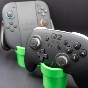 Może przedstawiać: Wyświetlane są dwa kontrolery Nintendo Switch, jeden szary i jeden czarny. Kontrolery spoczywają na jasnozielonych, cylindrycznych stojakach. Stojaki przypominają rury z serii gier wideo Super Mario.