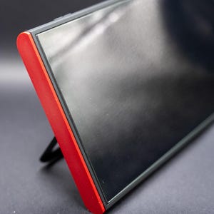 以下が含まれることがあります： 赤色のアクセントが付いた黒い電子機器。わずかに上向きに傾いています。デバイスは黒い画面と背面にスタンドが付いています。赤いアクセントはテクスチャ加工されています。背景は濃い灰色です。