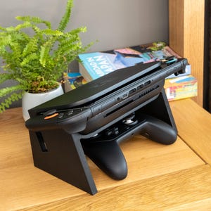 Puede incluir: Consola de juegos negra apoyada en un soporte de madera negro. La consola tiene detalles en naranja y está inclinada hacia arriba. Un mando es visible debajo. Un helecho en maceta y libros están en el fondo.