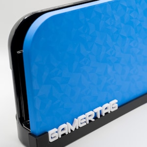 Puede incluir: Una cubierta azul para consola de juegos con un patrón geométrico, sujeta en un soporte negro. La palabra "GAMERTAG" se muestra en blanco en el soporte. La consola tiene un borde negro brillante.