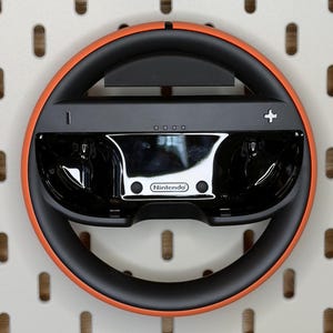 Può includere: Accessorio per volante Nintendo nero e arancione. Il volante ha un centro nero con un segno più e quattro piccoli punti. L'anello esterno è arancione. Il logo Nintendo è visibile sulla parte anteriore.