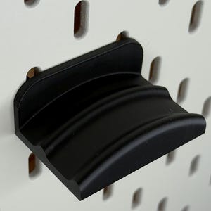 Peut inclure: Une étagère noire conçue pour un système de panneau perforé. L'étagère est en matériau solide et présente un bord avant incurvé. Elle est montée sur un panneau perforé blanc avec des trous ovales espacés.
