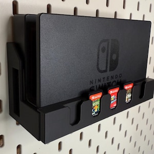 Puede incluir: Base negra para Nintendo Switch con ranuras para cartuchos de juego. La base sujeta la consola Switch y varios cartuchos de juego. El logotipo de Nintendo Switch es visible. La base está montada en una pared perforada blanca.