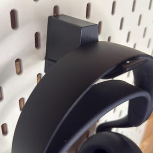 Peut inclure: Casque audio noir suspendu à un support mural noir. Le support est fixé à un panneau perforé blanc avec des trous ovales espacés régulièrement. Le casque est incurvé et possède de grands écouteurs.