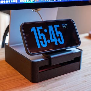 May include: A black digital clock showing the time 15:45 in blue numerals. The clock is mounted on a black and white stand, placed on a wooden surface. The date and temperature are also displayed on the clock.