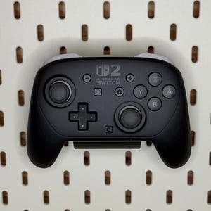 May include: A black Nintendo Switch Pro Controller with grey accents, featuring buttons and joysticks. The controller is displayed against a white pegboard background. The Nintendo Switch logo is visible.