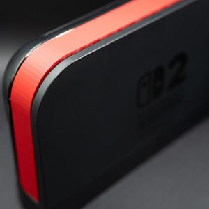 Könnte beinhalten: Nahaufnahme eines schwarzen elektronischen Geräts mit roter Akzentfarbe. Das Gerät hat abgerundete Kanten und eine glänzende Oberfläche. Der Text "Switch 2" ist sichtbar. Das Gesamtdesign ist elegant und modern.