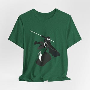 May include: A green t-shirt with a graphic of a character in a black cape and mask, holding a white lightsaber. The character is in a dynamic pose, with a black and white object below.